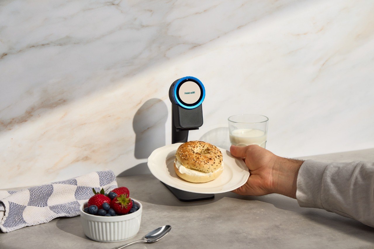 A bagel on a plate held up to the Trepo device on its countertop stand, the display reading Hold still